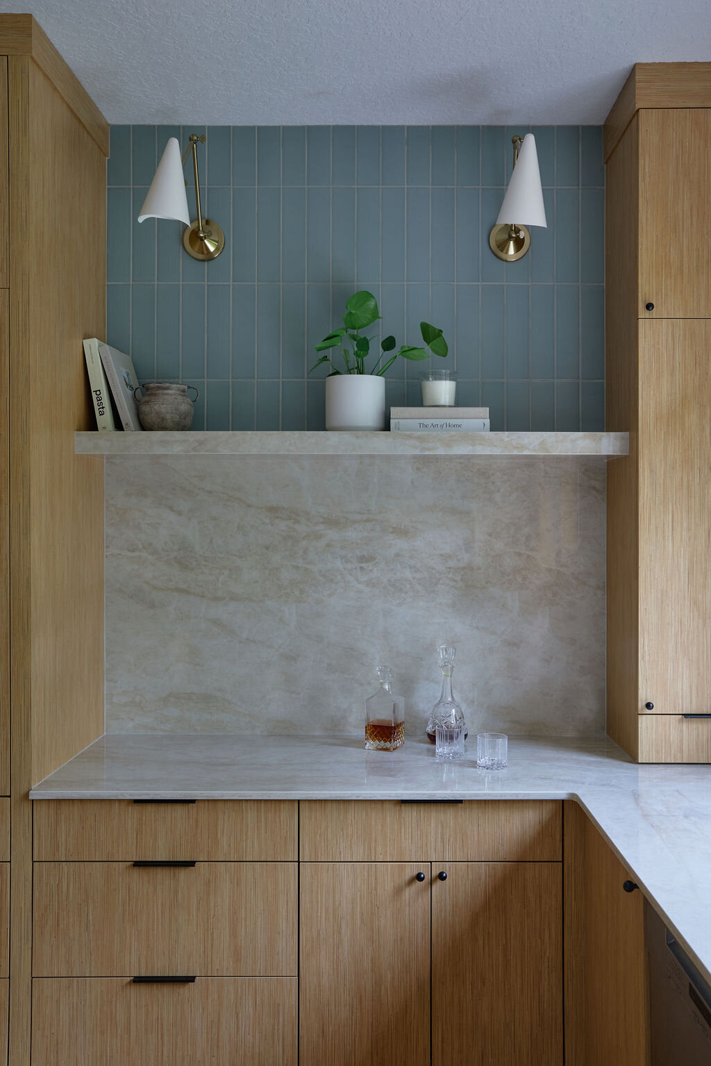 Kitchen with white oak cabinets, and slim simple black hardware. Taj mahal quartzite countertops that run up the backsplash to a quartz shelf with cookbooks and plants on it. Green subway tile backsplash above the shelf to the ceiling. Two sconces above the shelf that are modern, white and gold.