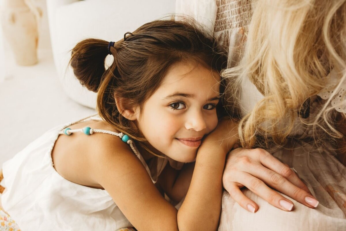 A little girl smiling with her brown hair tied up and leaning to her mother