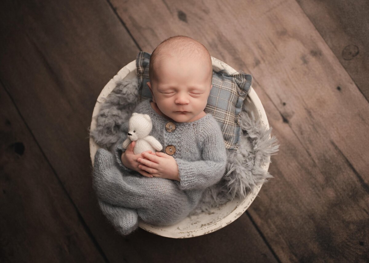 Aerial image. Baby boy in a grey knit onesie is laying in a basket with his legs folded atop of him. He is sleeping and holding a tiny felt teddy bear in his hands. Captured by best Murrieta newborn photographer Bonny Lynn Photography/