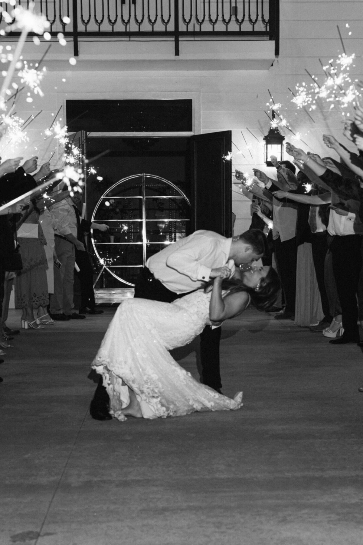 A black and white of the bride and groom doing a dip kiss while thier guests hold sparklers