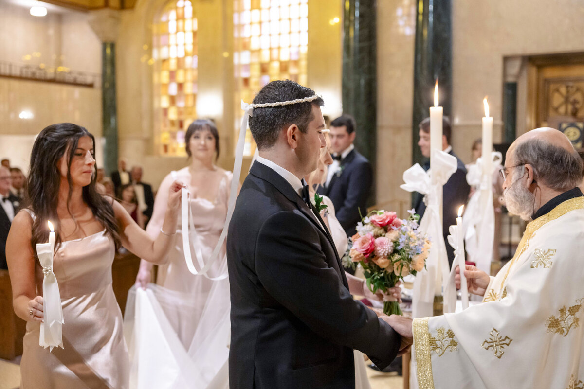 A man and woman stand in a church, wearing formal attire, and participate in a wedding ceremony. They are joined by a priest and two women holding candles. One woman also holds a bouquet of flowers. The church is filled with natural light.