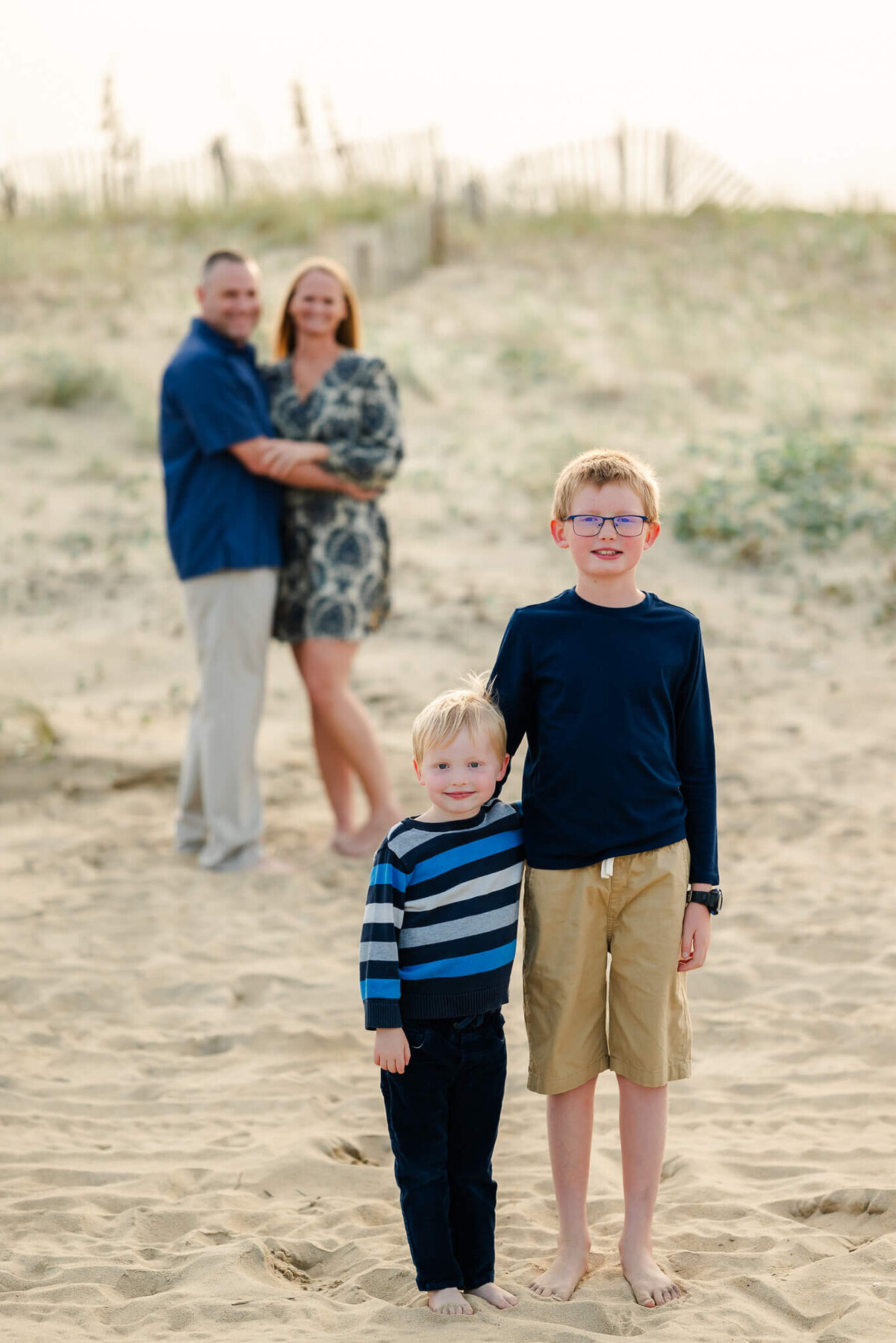Two brothers put their arms around each other while their parents do the same behind them at a family session on the beach.