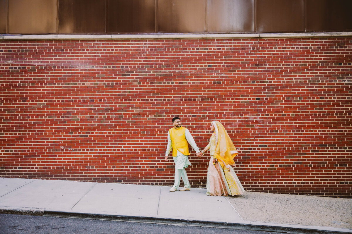 Bengali bride and groom wearing yellow outfits walking up a hilly street in front of a brick wall at their holud ceremony