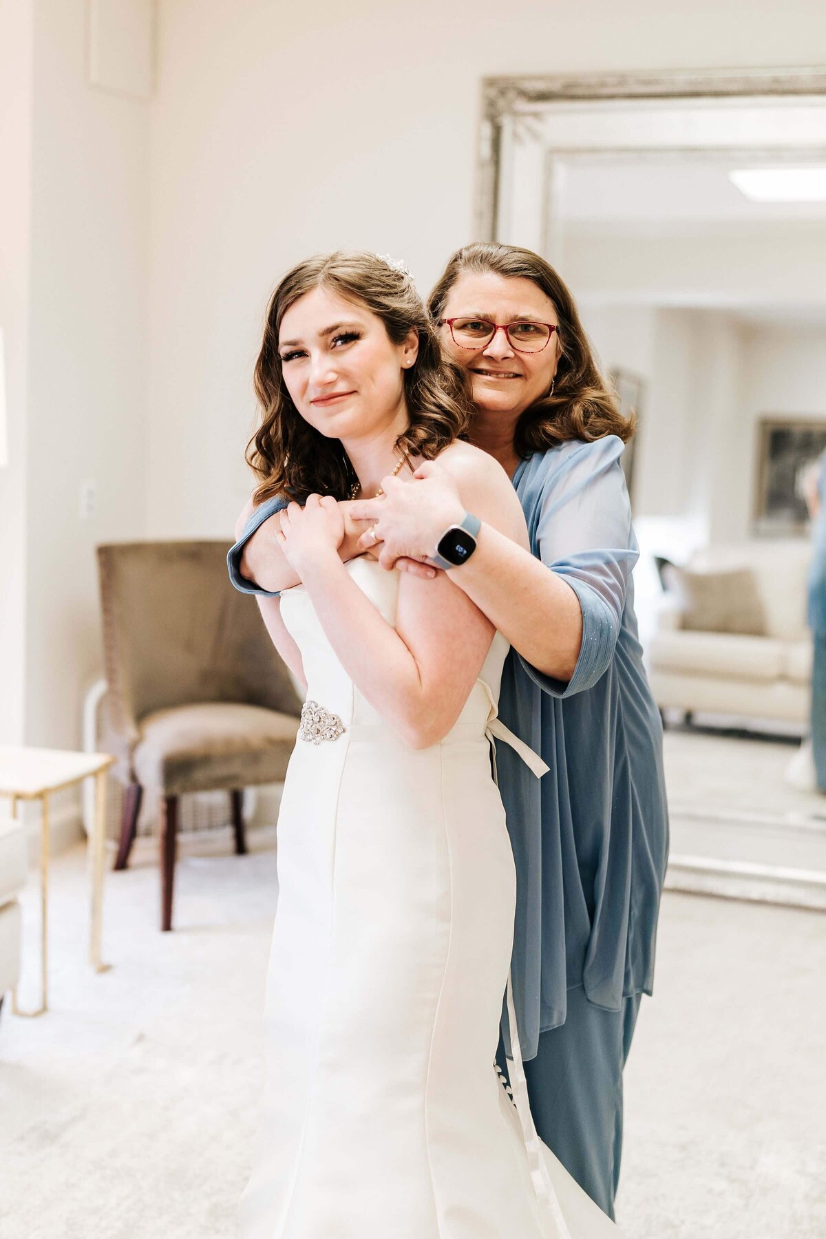 A bride in a white dress and a woman in a blue outfit embrace and smile warmly at each other in a bright bridal suite with a large mirror in the background.