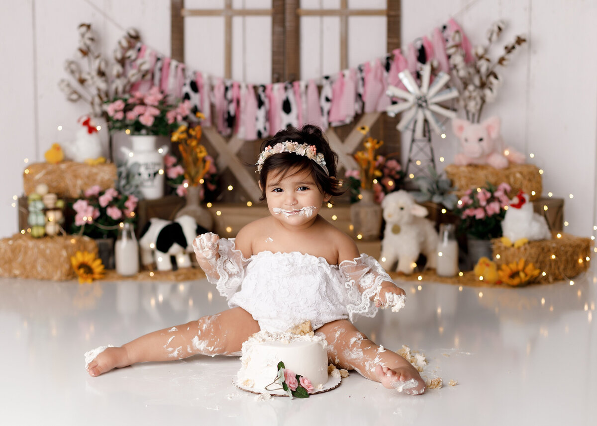 Pink cowgirl rustic barn cake smash in West Palm Beach and Boynton Beach newborn, cake smash, and portrait photography studio.Baby girl is wearing a white lace off the shoulder romper and flower crown with cake on her feet, legs, hands, and face. She is smiling at the camera.  In the background, there is a rustic barn door, bales of hay, stuffed farm animals, flowers, and a pink, white, and cow print banner.