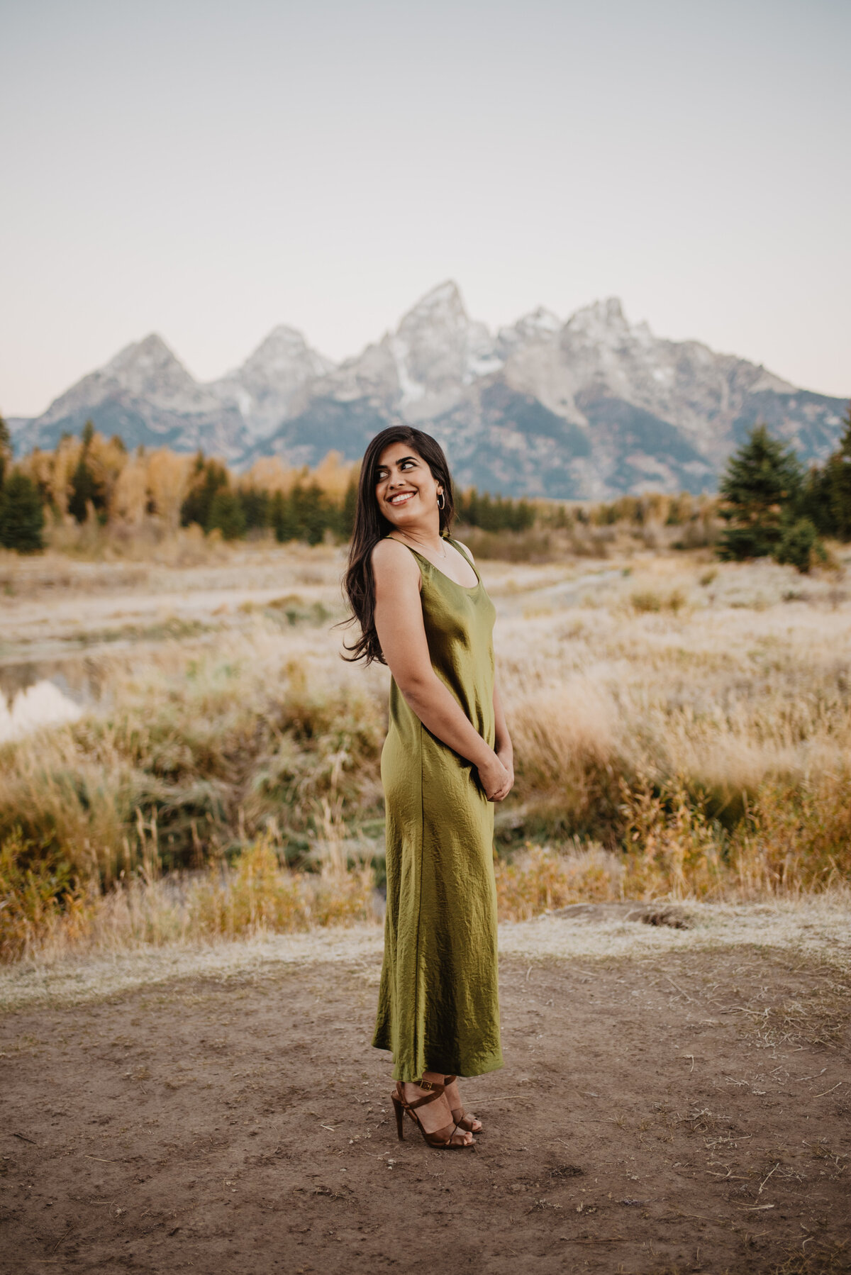 woman posing in an olive green dress in the fall in front of the Tetons while smiling and looking over her shoulder