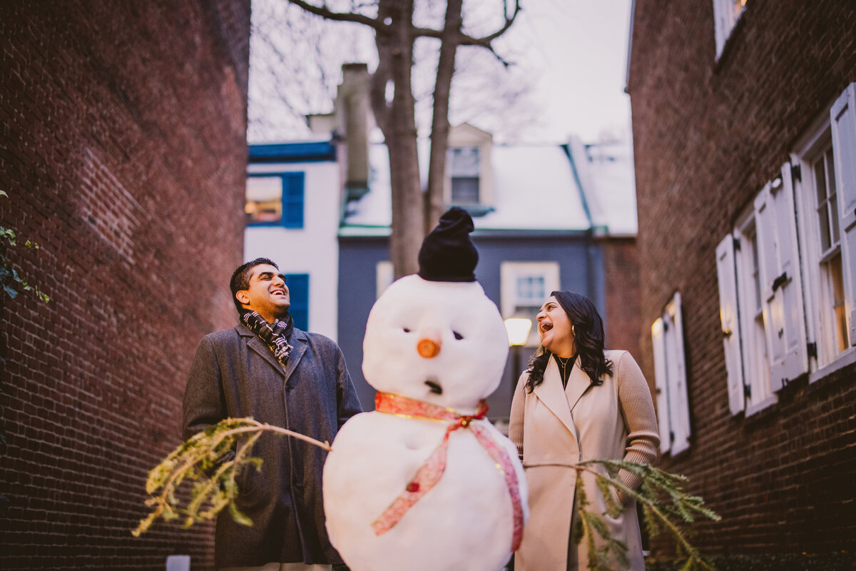 An engaged couple wearing coats laughing in between a snowman in the winter in downtown philadelphia