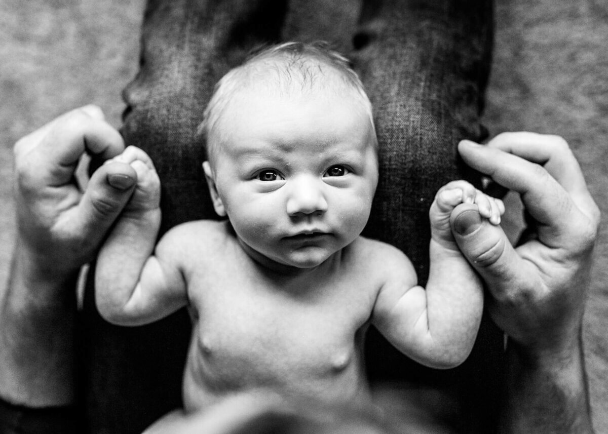 Portland newborn photographer takes black and white image of newborn baby laying in dads lap while holding both of his hands.