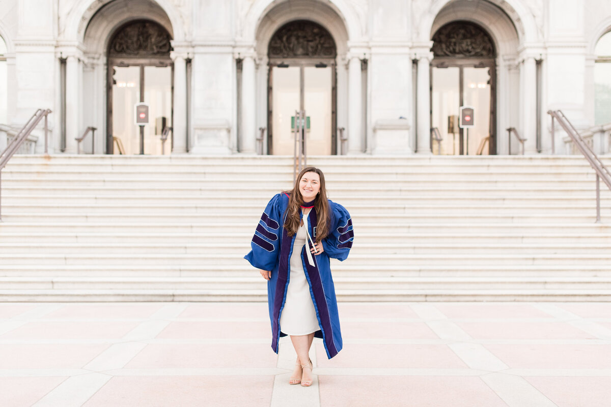 phd law grad blue gown hands on hips in front of marble staircase