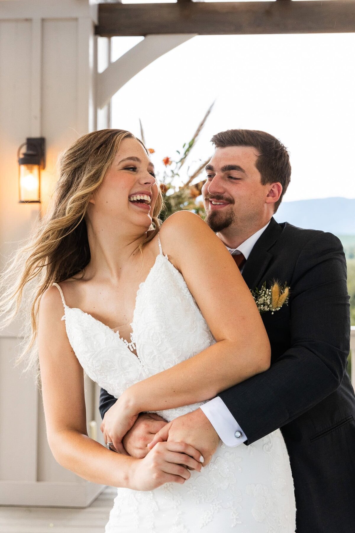 A groom playfully wraps his arms around his bride from behind as she throws her head back in laughter. The bride’s blonde waves dance in the wind as she smiles, dressed in a lace wedding gown, while her groom, wearing a charcoal suit, grins in the golden light of an outdoor venue
