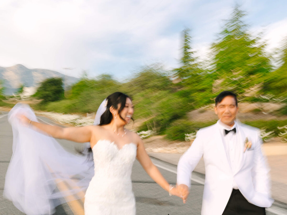 A joyful bride in a white gown and veil holds hands with a groom in a white tuxedo, both smiling and running. The background is a blurred road with greenery, conveying motion and happiness.