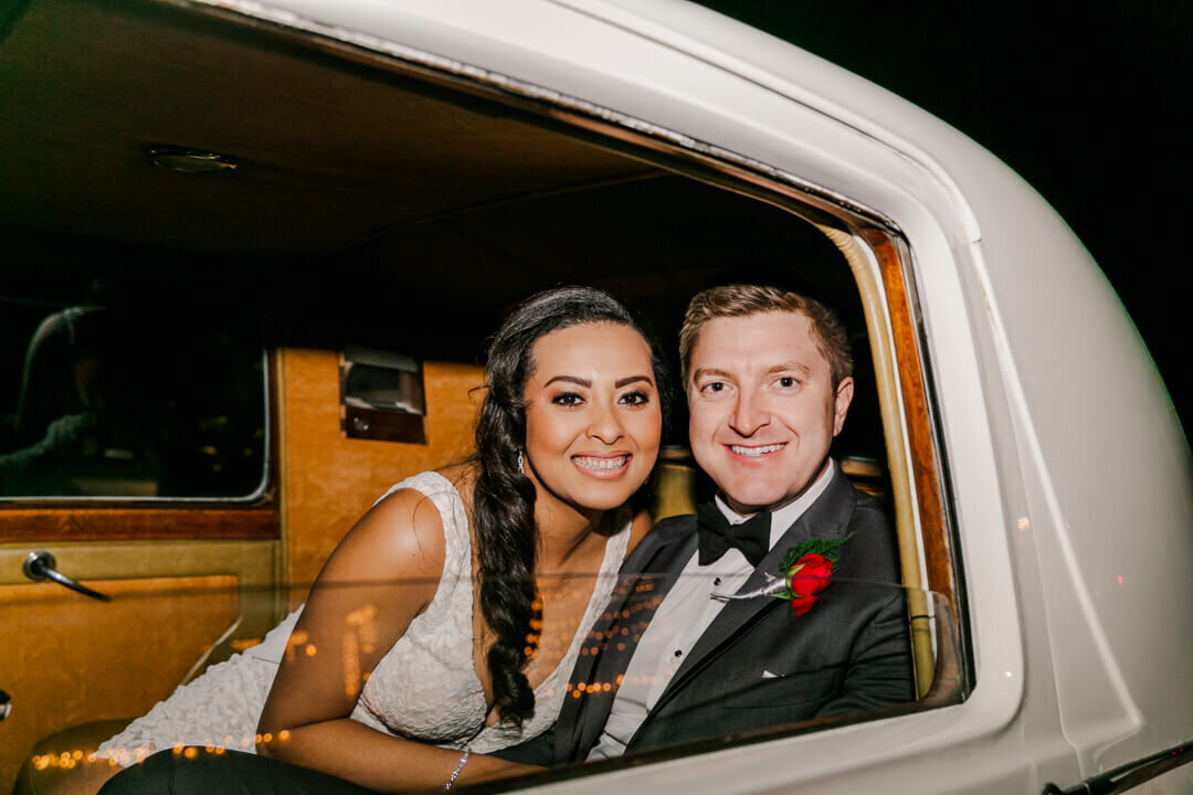 Newlyweds snuggle and smile out the window in the back of a white vintage car