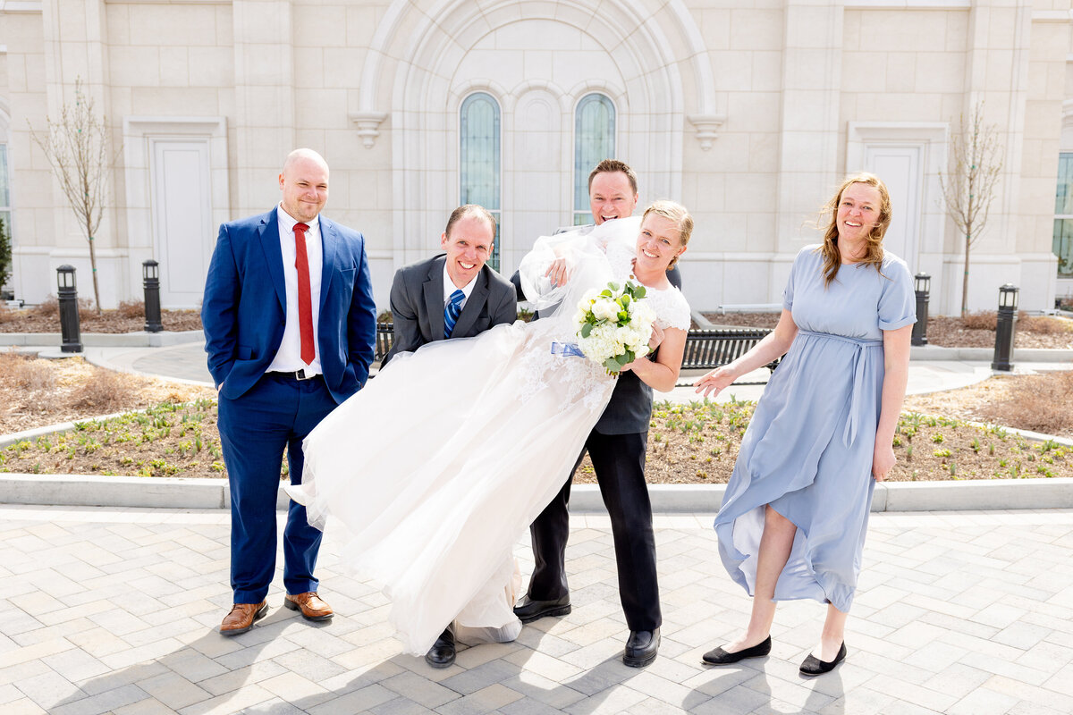 The bride is playfully lifted by her family members outside the LDS temple, all smiling and enjoying the moment.