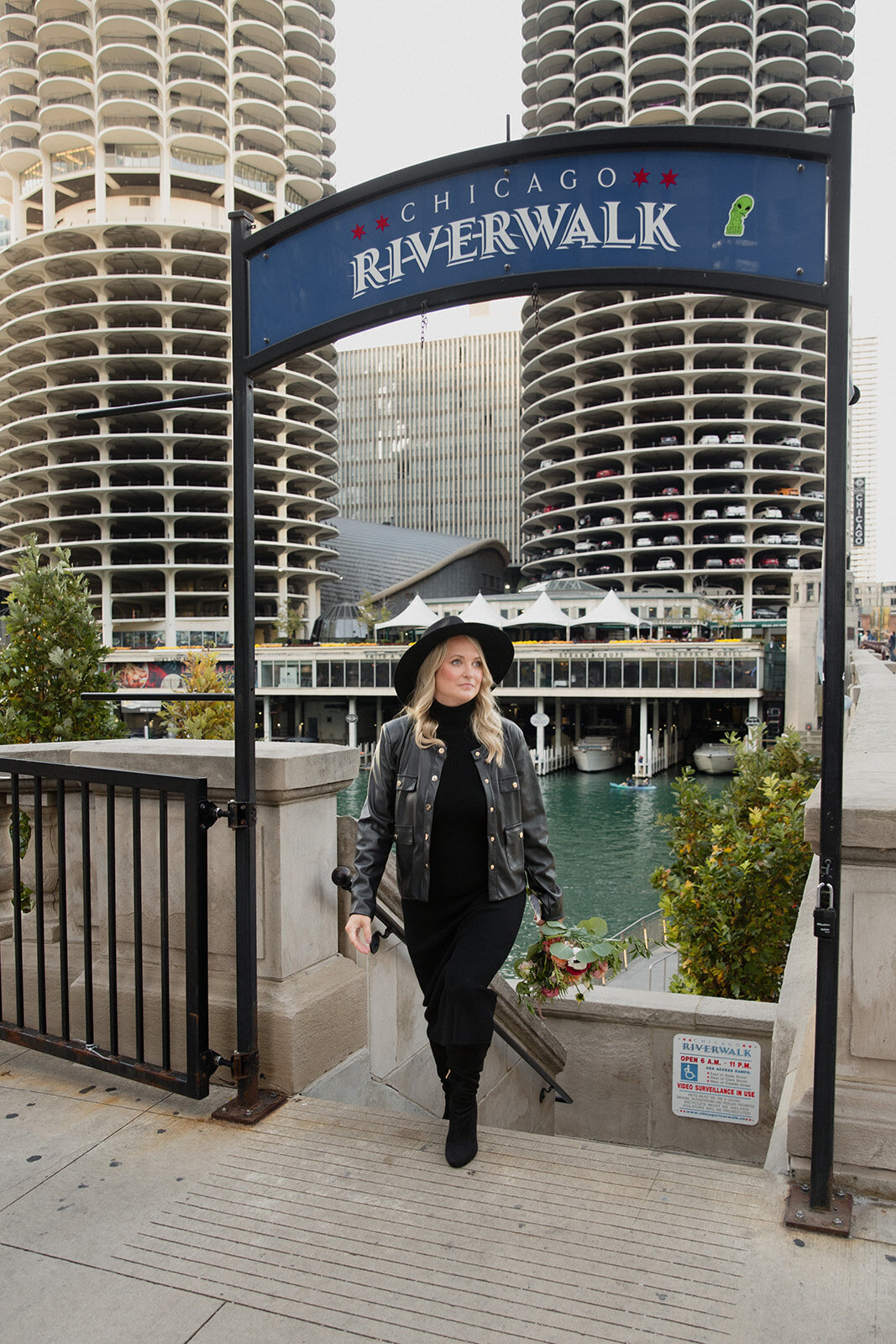 blonde woman in black dress, leather jacket, black boots and fedora is walking up the Chicago Riverwalk steps with river and buildings in background.