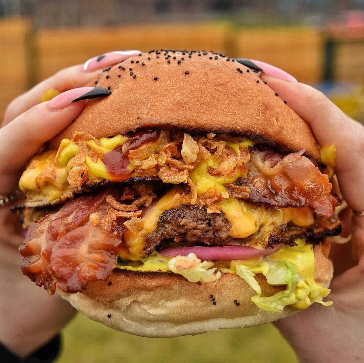 Double beef burger held in hands with cheese, bacon and lettuce hanging out. The hand has long painted nails