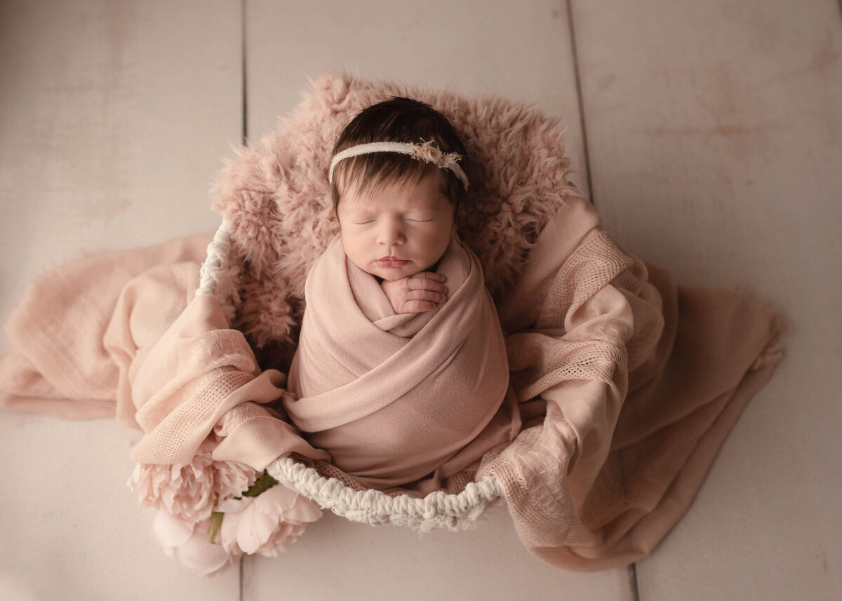 Baby Sleeping in basket with rose gold muslin draped across it. Baby in rose gold wrap with matching headband in Lake Elsinore photography studio. Aerial view.