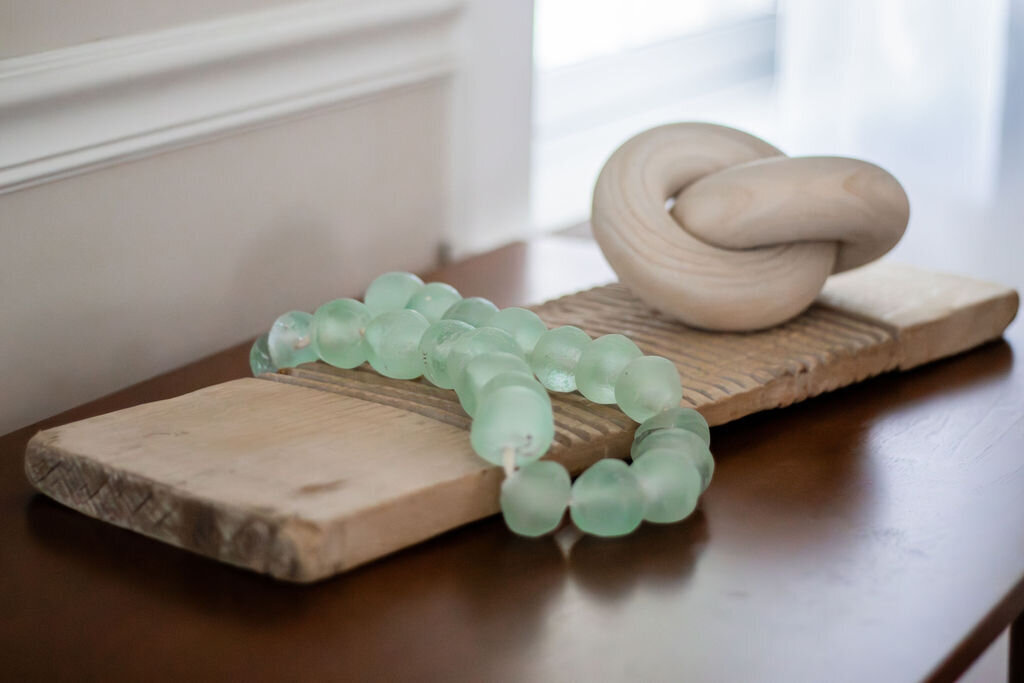 A close-up of a dark wooden tabletop displaying decorative objects. A strand of translucent, light green glass beads is draped across a weathered, light-colored wooden plank with subtle ridges. Behind the beads, a smooth, light-colored wooden decorative knot rests on the same plank. The background shows a light beige wall with a horizontal molding and a softly blurred window area. The lighting is gentle and diffused.