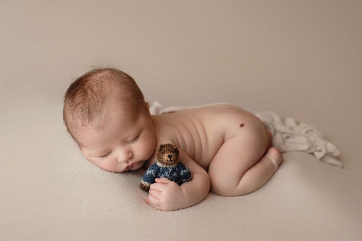 A newborn baby sleeps in froggy pose clutching a small stuffed bear in a New Orleans Newborn Photographer studio