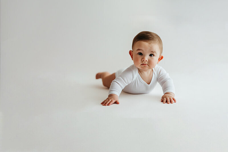 six month old baby boy in a tight white onesie laying on his belly on a white backdrop