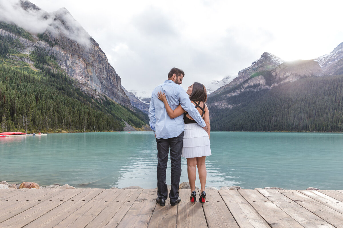 Engaged couple hold each other by the turquoise waters of Lake Louise, framed by the dramatic peaks of the Lake Louise glacier.