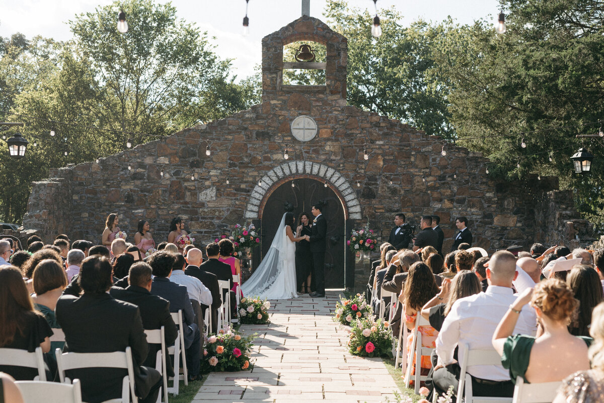 Wide ceremony view of an outdoor wedding at a rustic stone chapel, featuring lush aisle floral arrangements with roses, dahlias, and greenery framing the walkway as the couple exchanges vows surrounded by guests.
