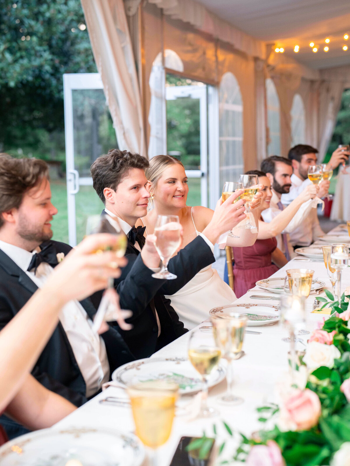 A joyful wedding reception with guests seated at a long table, holding glasses and toasting. The bride and groom are at the center, surrounded by friends and family. The table is elegantly set, and the tent is decorated with soft lights.