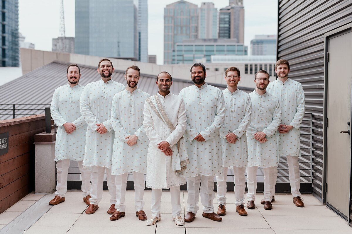 Hindu groomsmen wearing light blue sherwani with gold accents and white pants stand on either side of the groom who is wearing a white sherwani with a light blue and gold dupattan as they stand in front of the Nashville skyline at the Country Music Hall of Fame