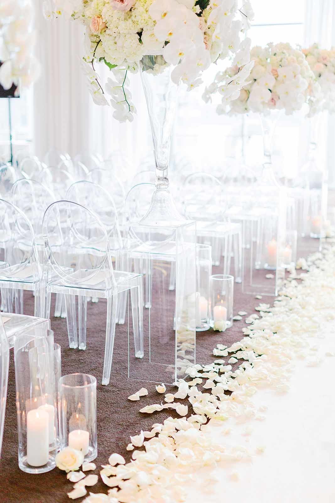 wedding ceremony aisle with large white flower arrangements, hurricane candles, white rose petals, white aisle runner, and clear lucite chairs