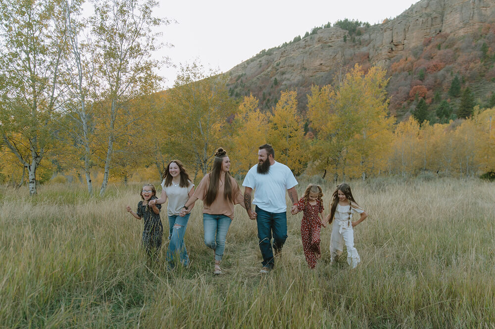 A family consisting of a mom, dad, and 4 daughters all walk towards the camera holding hands. They're all looking at one another and smiling.
