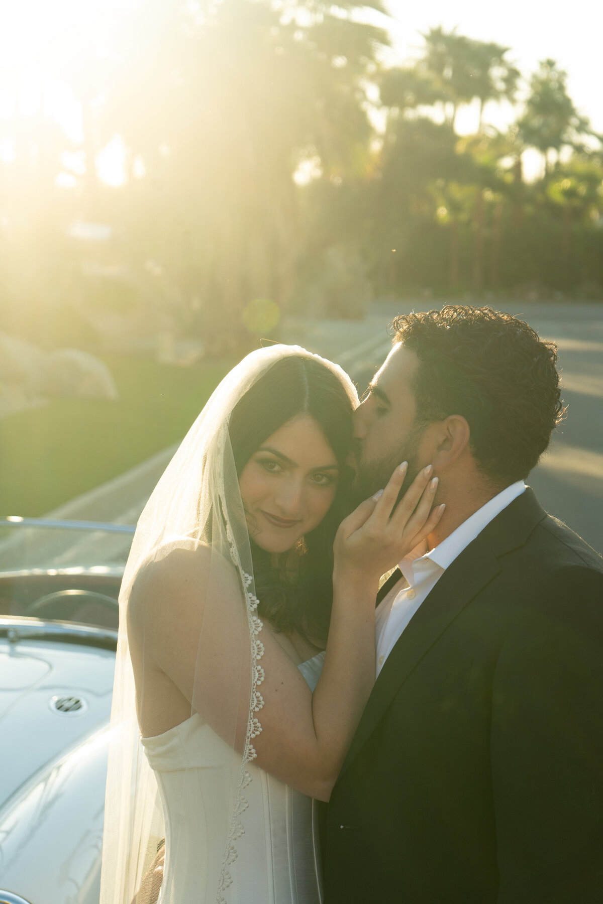 Portrait of bride and groom at sunrise in Palm Springs with her veil catching the glow of the sun and the groom kissing the top of her head