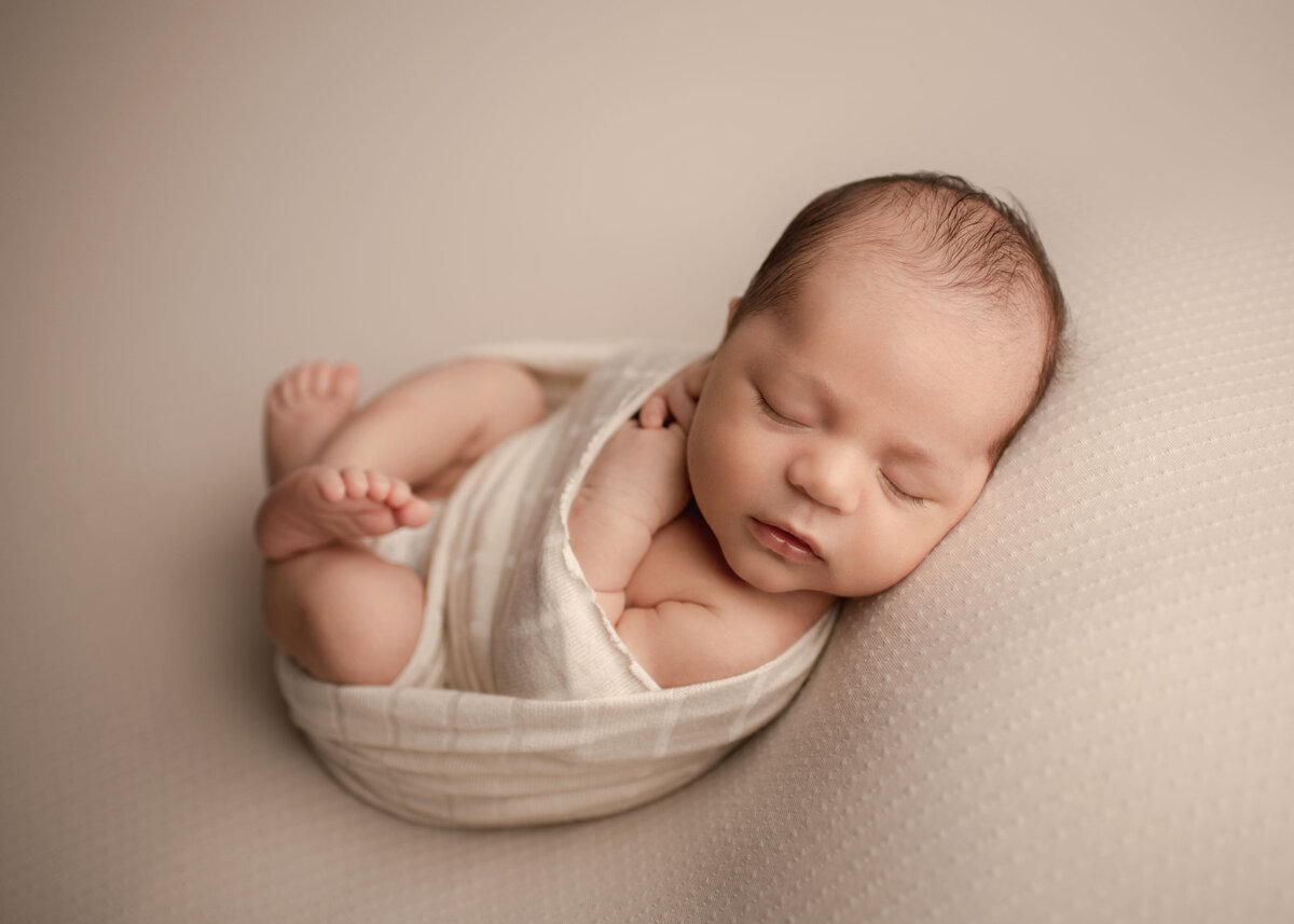 Lake Elsinore newborn photoshoot. Baby swaddled in a cream wrap with his legs peeking out and folded on top of him. His head is positioned toward the camera. Captured by best Lake Elsinore newborn photographer Bonny Lynn Photography.
