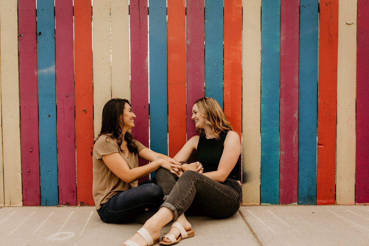 lesbian couple sitting and laughing in front of a rainbow fence