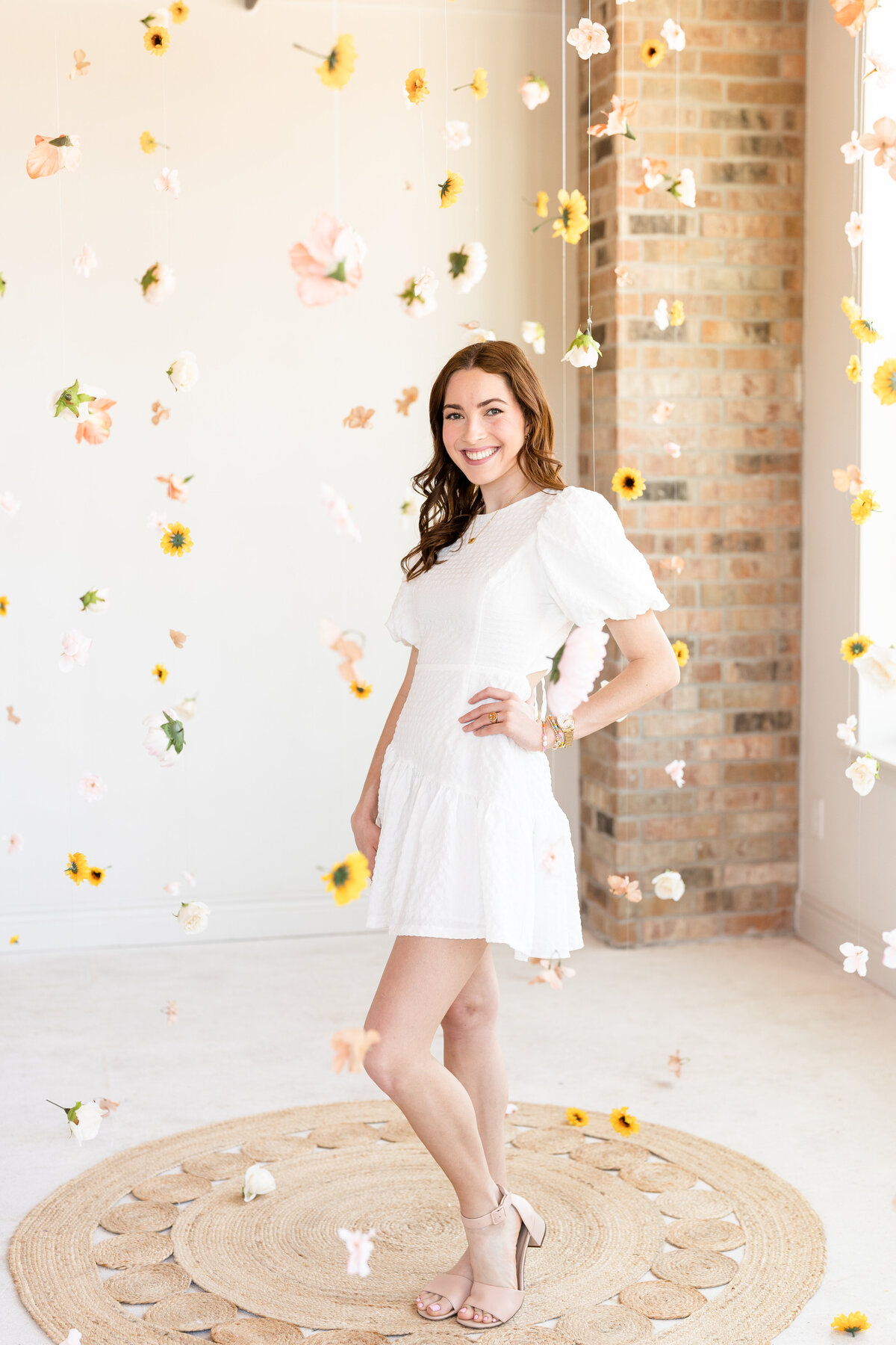 Texas A&M senior girl smiling with hand on hip while wearing white dress and in the middle of hanging flowers at Bravely Studio