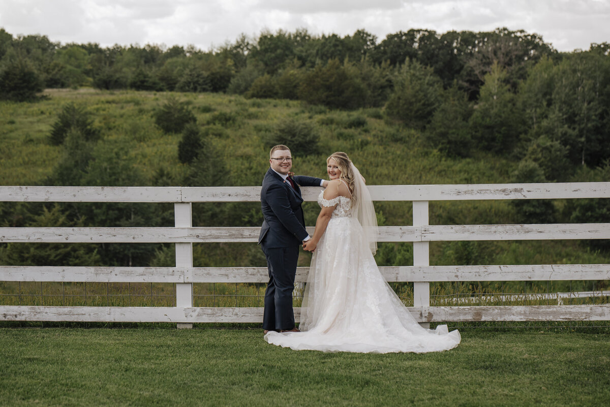 A romantic moment captured with a bride and groom standing by a fence in a picturesque countryside setting, sharing a glance and smiles on their wedding day taken by jen Jarmuzek photography a Minneapolis wedding photographer