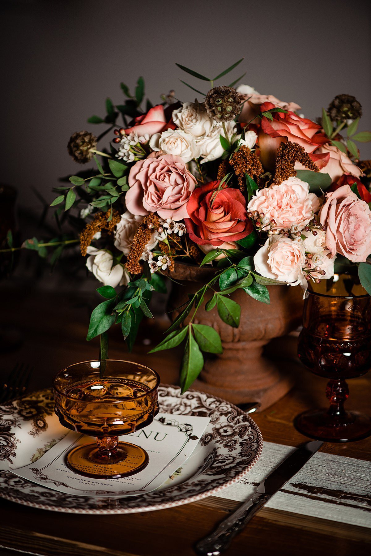 A farm table set with a floral white and brown plate with a menu and brown glass dessert bowl in the center. Silverware and an amber water glass  are on either side of the plate. Above the plate on the farm table is a large floral centerpiece in a brown footed compote bowl with blush roses, blush carnations, orange roses, white garden roses, brown flowers and greenery.