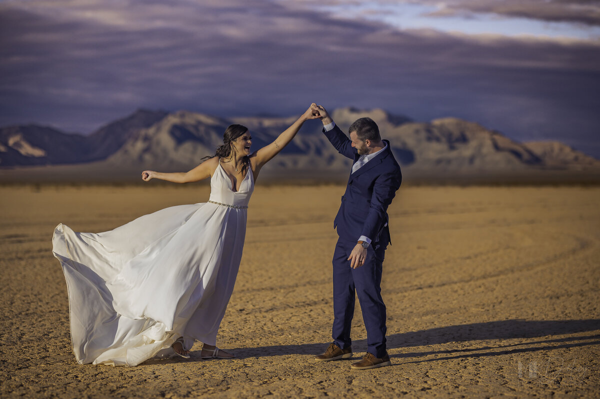 Dry lake Bed elopement Blue Suit on Groom  flowers by michelle  bride in cream color wedding dress with deep  plunging  neckline mountain skyline  sunset las vegas wedding photographers mk delacy photography