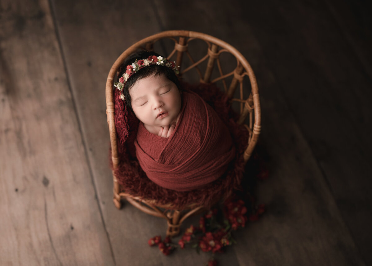 Aerial image of Riverside, CA newborn photoshoot. Baby girl wrapped in rust stretch fabric and sleeping on a newborn posing prop chair. Captured by Best Riverside, CA newborn photographer Bonny Lynn Photography.