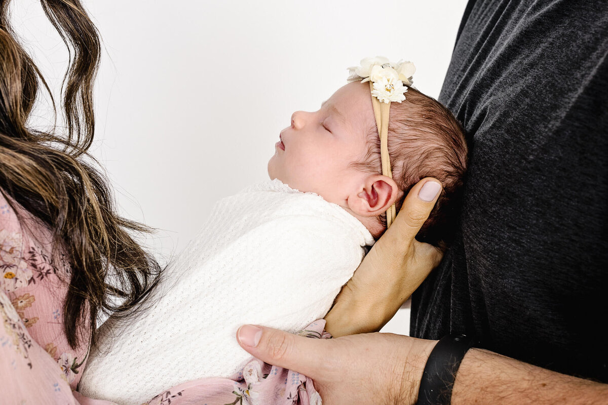 A baby with a floral headband sleeps peacefully, wrapped in a white blanket. The baby is held by a person wearing a dark shirt, with another person in a floral outfit nearby. The background is a plain white.