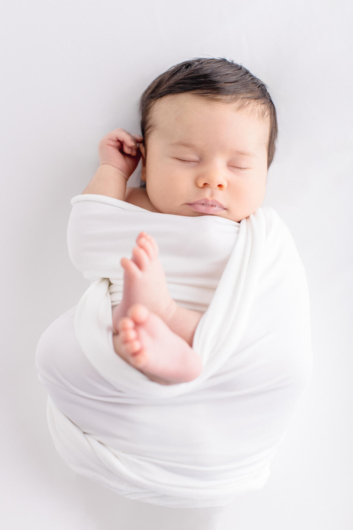 A peacefully sleeping baby swaddled in a soft white blanket lies on a white surface. The babys eyes are closed, and their small hand is near their head, with tiny feet visible peeking from the wrap.