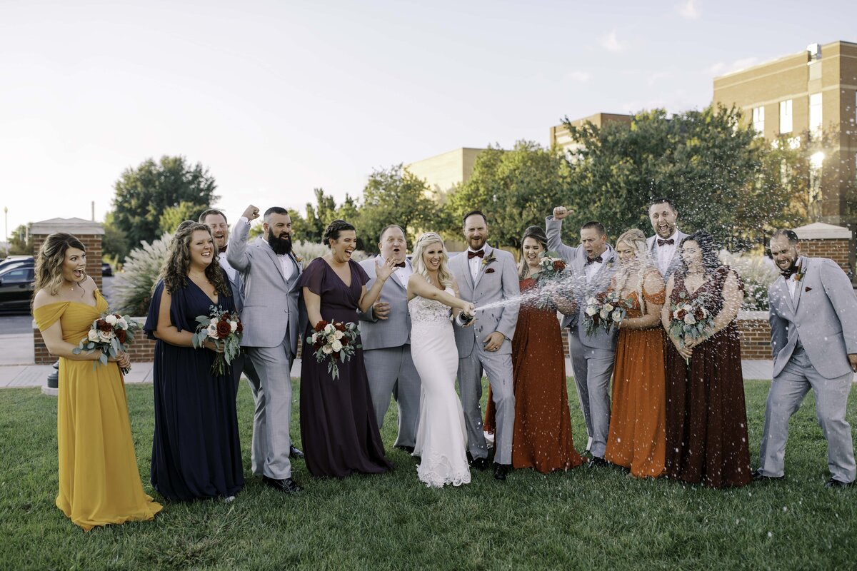 A wedding party with the bride and groom stand together in a semi circle popping a bottle of champagne and laughing together