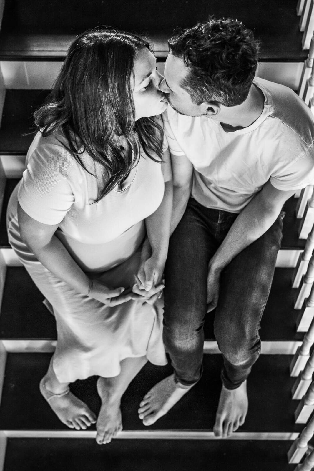 A black and white image, shot from above, of an expecting couple kissing on their staircase.