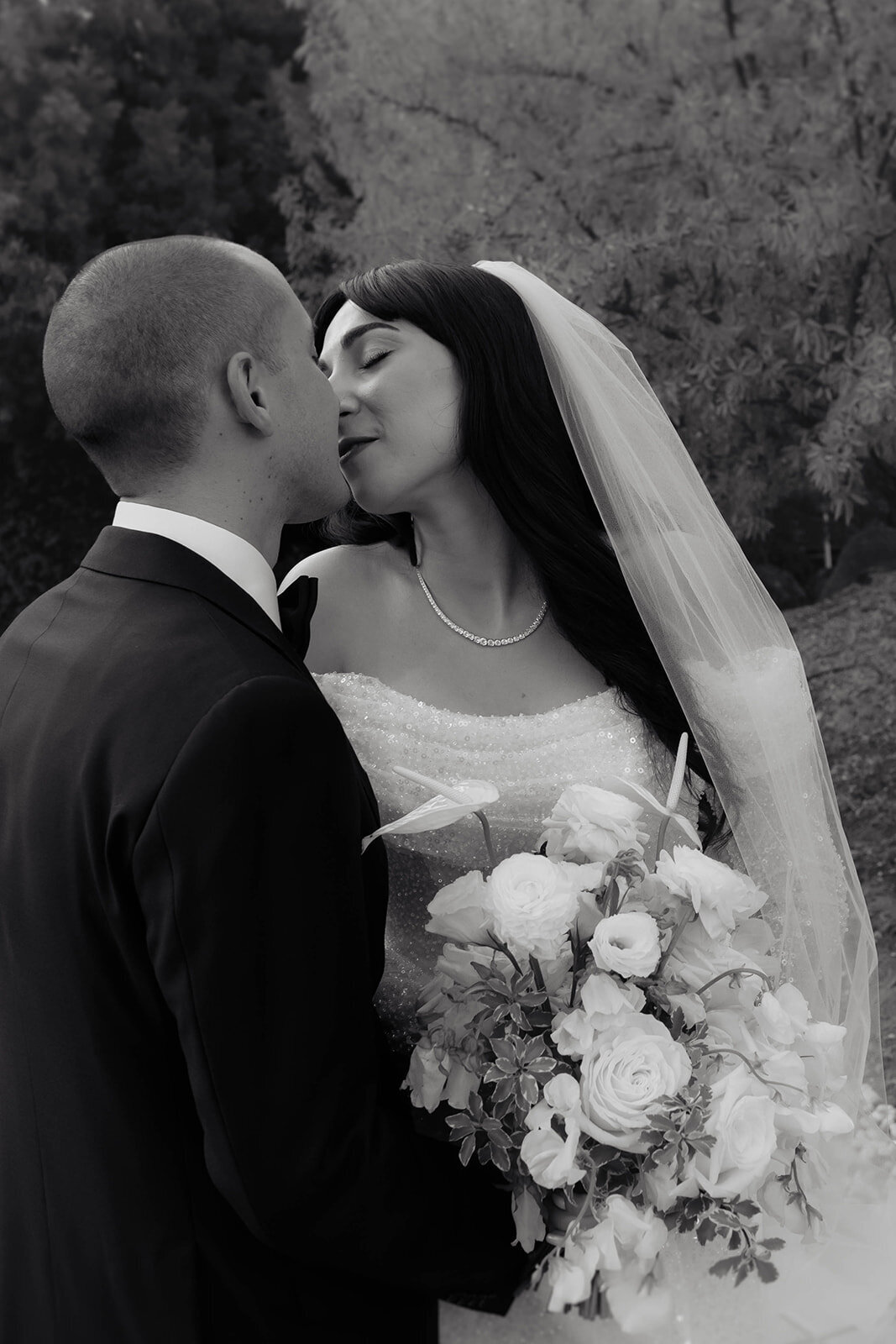 A black and white photograph of a bride and groom kissing, holding a bouquet of white roses.