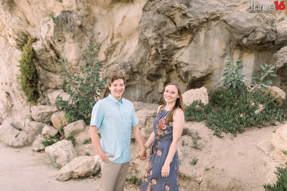 Engaged couple pose in front of a large cliff holding hands