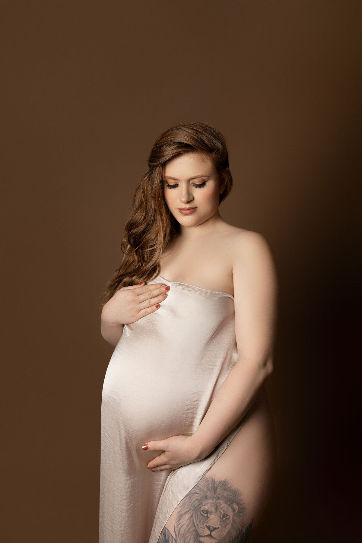 Maternity photos in top London, Ontario studio. Ogg Photography captures woman with long red hair sweeping over her shoulder. The woman is draped in cream-coloured silk fabric with one hand over her breast and the other under her baby bump. Her front leg is exposed bearing a lion tattoo.