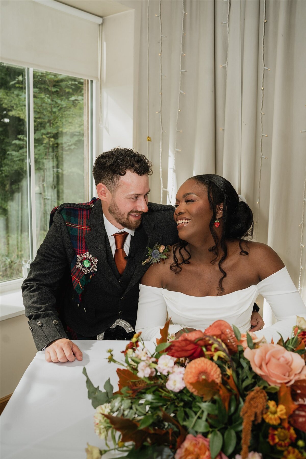 A bride and groom smile candidly at each others they sign their marriage licence. A large bouquet sits in the foreground of the photograph.