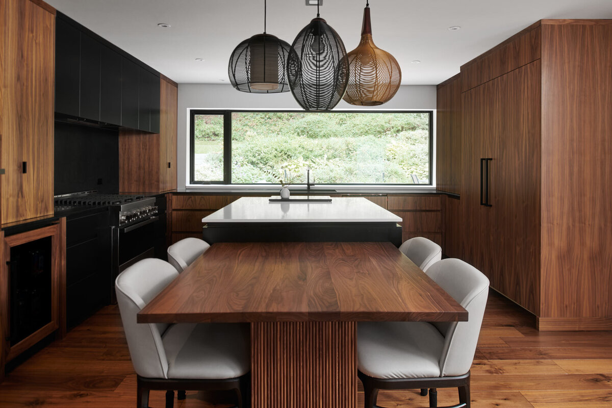 A modern kitchen in Richmond Hill, Ontario, captured by a professional interior design photographer specializing in projects across Richmond Hill, Toronto, and the Greater Toronto Area. The space features custom walnut cabinetry, a wooden dining table with upholstered chairs, unique pendant lighting, and a white countertop island overlooking a lush green backyard through a wide window.