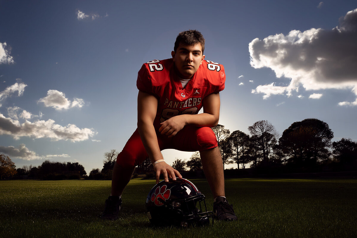 Senior football player in vibrant red uniform, crouched in a powerful stance on the field. The dramatic sunset backdrop adds to the dynamic energy, perfect for Dallas senior photography focused on sports and action.