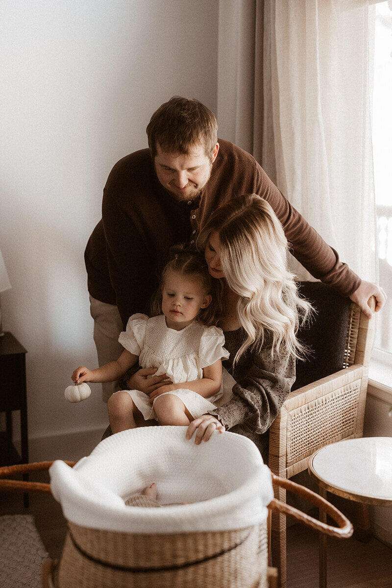 of three cuddle near a window while looking down at their new baby in a Moses basket