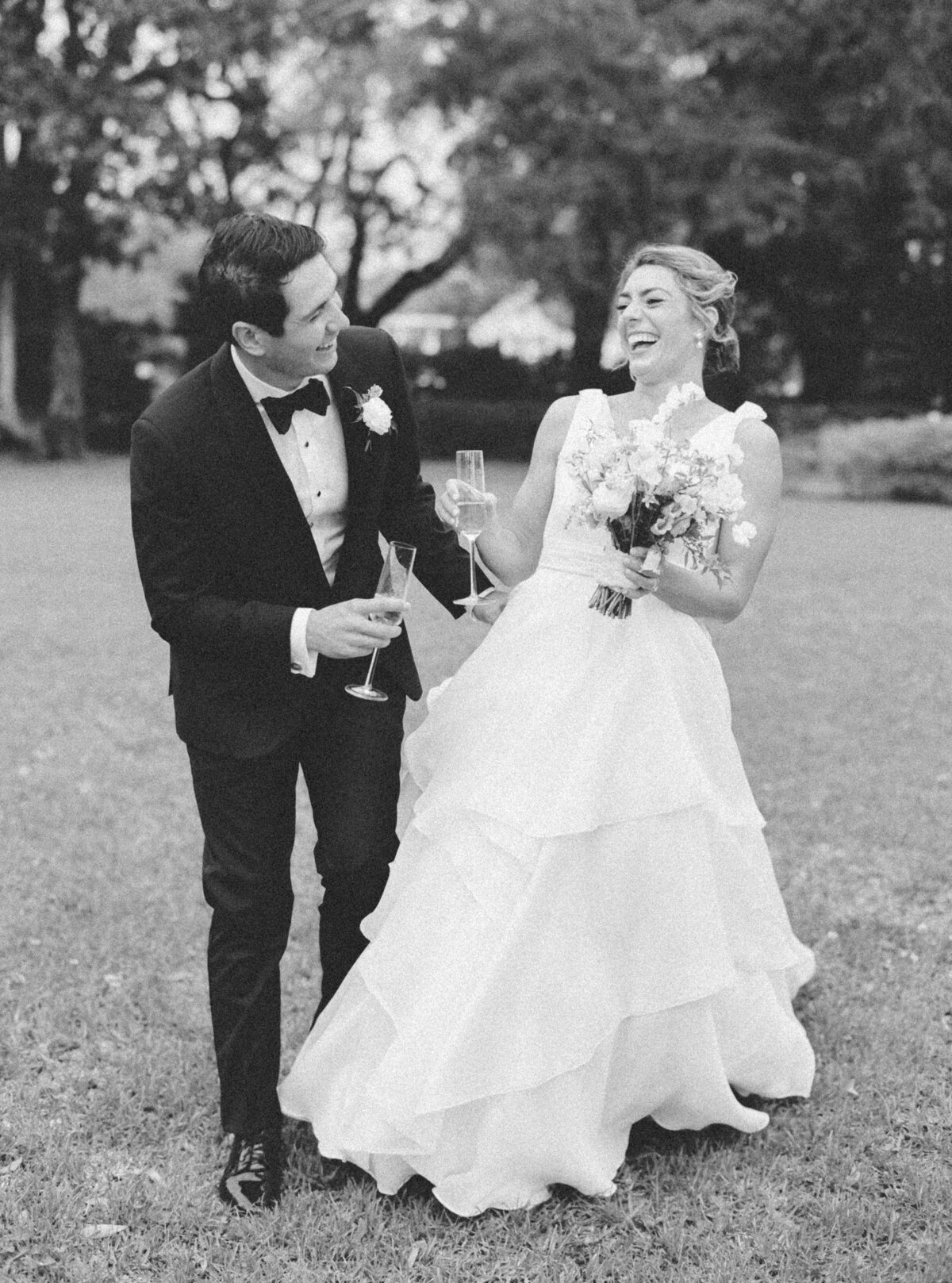 Black and white film photographer. Bride and groom laugh while sipping champagne on the front lawn at Lowndes Grove during cocktail hour. Destination wedding photographer. Kailee DiMeglio Photography.