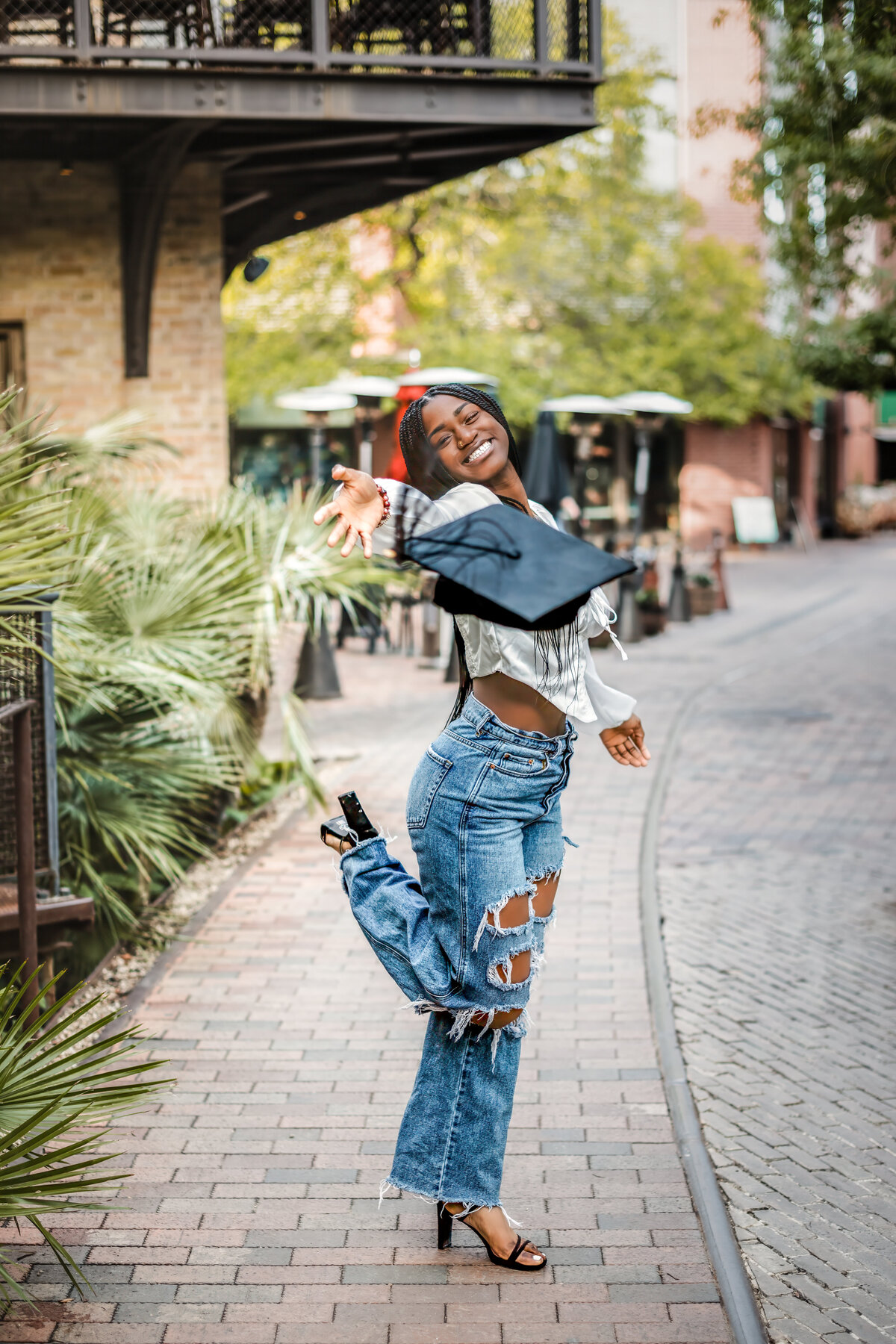 Steel High School senior girl tossing graduation cap at The Pearl in San Antonio, wearing jeans and smiling.